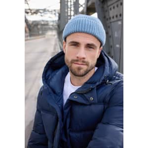A man wearing a blue fisherman beanie and a navy puffer jacket stands outdoors with a bridge in the background.