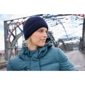 A navy blue knitted winter beanie worn by a person in a teal puffer jacket, set against an industrial bridge backdrop.