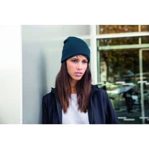 A knitted beanie in dark green worn by a woman with long hair, standing against a modern backdrop.