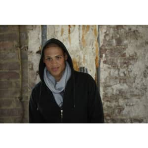 A person wearing a grey fleece scarf and a black hoodie stands against a weathered brick wall.