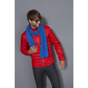 A man in a red jacket wears a blue scarf. He is smiling and posing against a grey background.