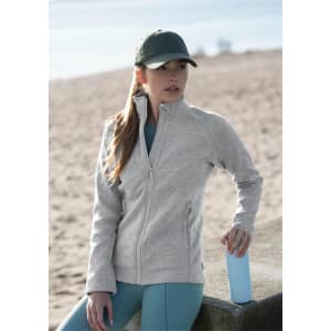 A woman wearing a light grey polar fleece jacket and a cap stands by a sandy area, holding a blue drink bottle.