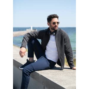 A men's polar fleece jacket in dark grey, worn by a man sitting by the sea, paired with navy pants and sunglasses.