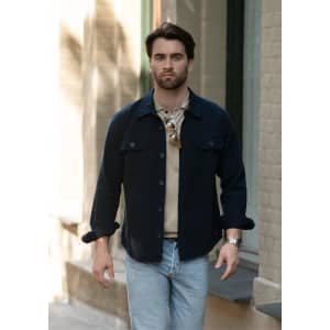 A unisex shirt in navy with buttons, worn over a beige top, paired with light jeans. The model is walking outdoors.