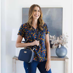 A smiling woman wearing a floral navy top holds a small navy bag, standing beside a decorative vase and artwork.