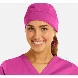 A unisex scrub cap in bright pink, worn by a smiling woman in a matching scrub top. The cap features a logo.