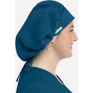 A unisex bouffant cap in deep teal, featuring a logo, worn by a model showcasing its profile.