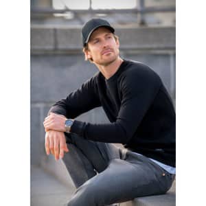 A black sweatshirt with long sleeves worn by a man sitting on stairs, paired with grey jeans and a cap.