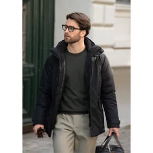 A man in a black jacket walks outside, wearing glasses and carrying a bag, with a green top and light trousers.