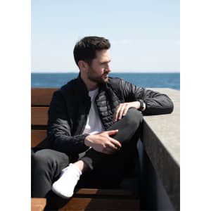 A puffer jacket in black, worn by a man seated on a bench with a seaside backdrop. The jacket features a logo.