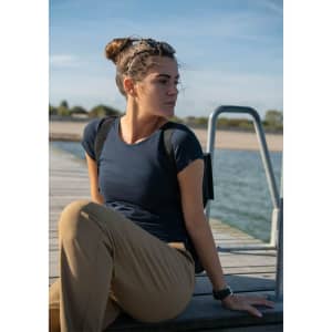 A woman sits on a dock wearing a navy blue T-shirt and beige trousers, with a serene water background.