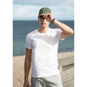 A white men's T-shirt displayed outdoors, with a model wearing a green cap and grey shorts by the beach.