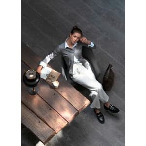 A woman in a grey cardigan sits on a wooden table, wearing light trousers and black shoes, with a lamp and notebook nearby.