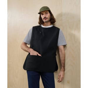 A black Utility 2.0 Vest Apron with pockets is worn by a model in a striped shirt against a wooden background.