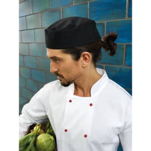 A black chef's hat is worn by a man in a white chef's coat, holding a basket of vegetables.