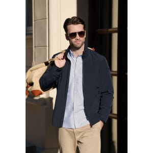 A navy jacket is worn by a man with sunglasses, holding a backpack, standing near a building.