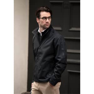 A man wearing a black jacket stands outdoors, paired with light trousers and glasses, against a dark background.