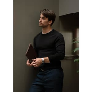 A black knitted jumper worn by a man leaning against a wall, holding a brown folder in his hand.