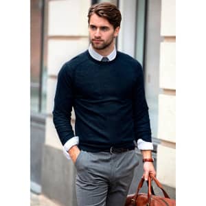 A man wearing a navy knitted jumper over a collared shirt, paired with grey trousers and holding a brown leather bag.