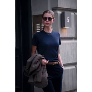 A woman wearing a navy blue t-shirt, holding a brown jacket and wearing sunglasses, stands in front of a building.