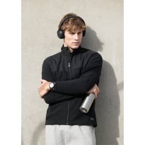 A black men's jacket is worn by a model holding a drink bottle, with headphones on and posing against a grey wall.