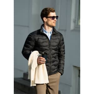 A black puffer jacket worn by a man holding a drink, with sunglasses and a white jacket draped over his arm.
