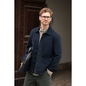 A man wearing a navy unisex shirt, paired with a light grey sweater and green trousers, holding a black bag.