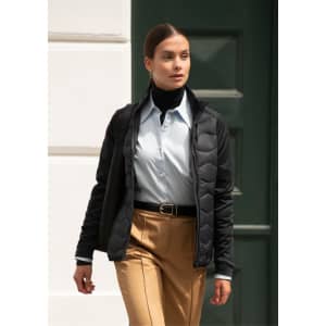 A woman wearing a black puffer jacket over a light blue shirt and tan trousers, standing against a green door.