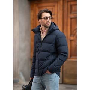 A man in a navy blue puffer jacket with a hood, wearing sunglasses and holding a bag.