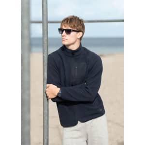 A man wearing a navy polar fleece jacket and sunglasses stands by a beach, leaning against a metal pole.