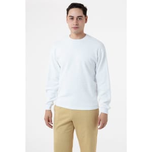A man wearing a white crewneck sweater, styled with beige trousers. The sweater features long sleeves and has a logo.