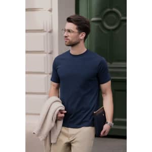 A navy men’s t-shirt is worn by a man holding a beige sweater and a clutch, set against a green door backdrop.