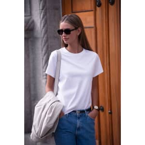 A white women's t-shirt is worn by a model with sunglasses against a wooden background, carrying a light-coloured bag.