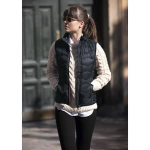 A woman wearing a black puffer vest over a cream knitted sweater, standing outdoors with hands in pockets.