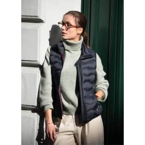 A navy vest is worn over a light green sweater, paired with cream trousers. The model has glasses and is leaning against a wall.