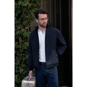 A man wears a navy hybrid jacket, holding a silver suitcase, standing by an ivy-covered wall. The jacket has a logo.