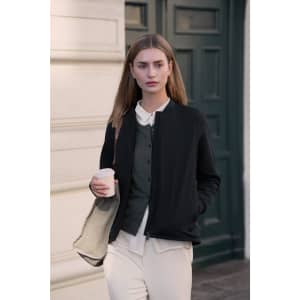 A woman in a black hybrid jacket holds a coffee cup, standing in front of a green door, wearing neutral-toned trousers.