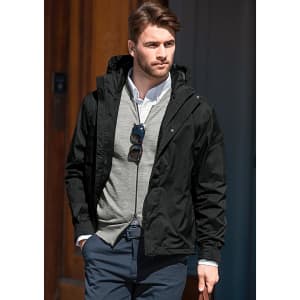 A man wearing a black soft shell jacket over a grey sweater, styled with sunglasses and smart trousers.