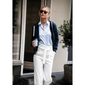 A woman wearing a navy blue hoodie over a light blue shirt and white trousers, holding a grey drink cup.