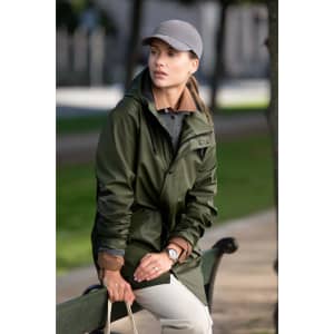 A green unisex jacket is worn by a woman sitting on a bench, paired with a grey cap and neutral trousers.