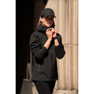 A black women's shell jacket is worn by a model, paired with a matching cap, standing against a textured wall.