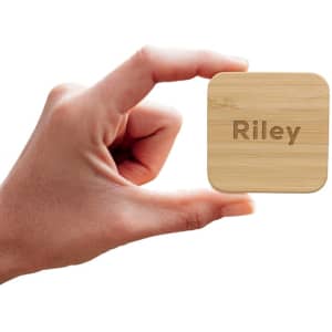 A hand holding a square bamboo wireless charger with the name "Riley" engraved on it.