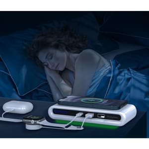 A wireless charging station in white and green, charging devices beside a sleeping person in a dimly lit bedroom.