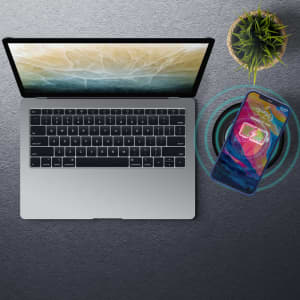 A silver laptop is placed on a textured surface next to a phone wirelessly charging. A small plant is also visible.