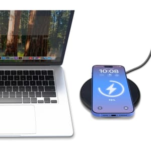 A black wireless charger with a smartphone displaying a charging indicator, beside a laptop with a natural backdrop.