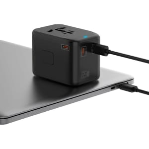 A compact black travel adaptor with multiple ports sits on a laptop, featuring a logo and connected cables.