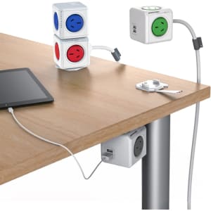 A Power Cube Extended Duo USB in white with red, blue, and green sockets, positioned on a wooden table alongside a tablet.