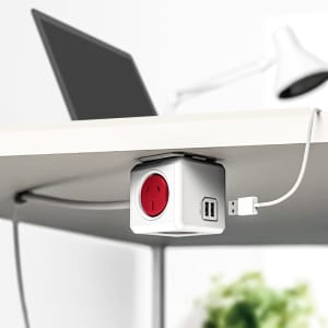 A white power cube with a red socket and two USB ports, mounted under a desk near a laptop and a lamp.