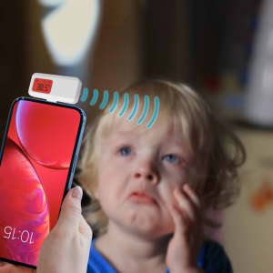 A smartphone with a white thermometer attachment displays a temperature reading near a child with a distressed expression.