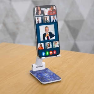 A white foldable phone stand with a blue patterned base, holding a smartphone displaying a video call.